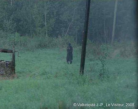 Le Visiteur, photogramme 26, plan 54, A travers l'obscurité de la forêt, sous une pluie battante, surgit un homme habillée sombrement, comme s'il annonçait un sombre avenir. Il est habillé d'un long manteau et un chapeau noirs.